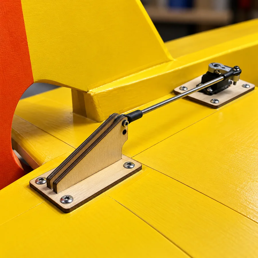 A laser-cut plywood control horn and servo mounting plate installed on the tail of a yellow 20-foot RC airplane, joined by a threaded pushrod